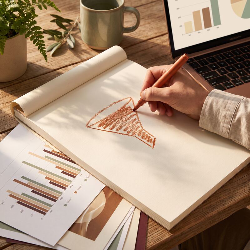 Hand sketching a PPC keyword-intent funnel on paper beside printed ad performance charts in warm afternoon light Hand sketching a PPC keyword-intent funnel on paper beside printed ad performance charts in warm afternoon light